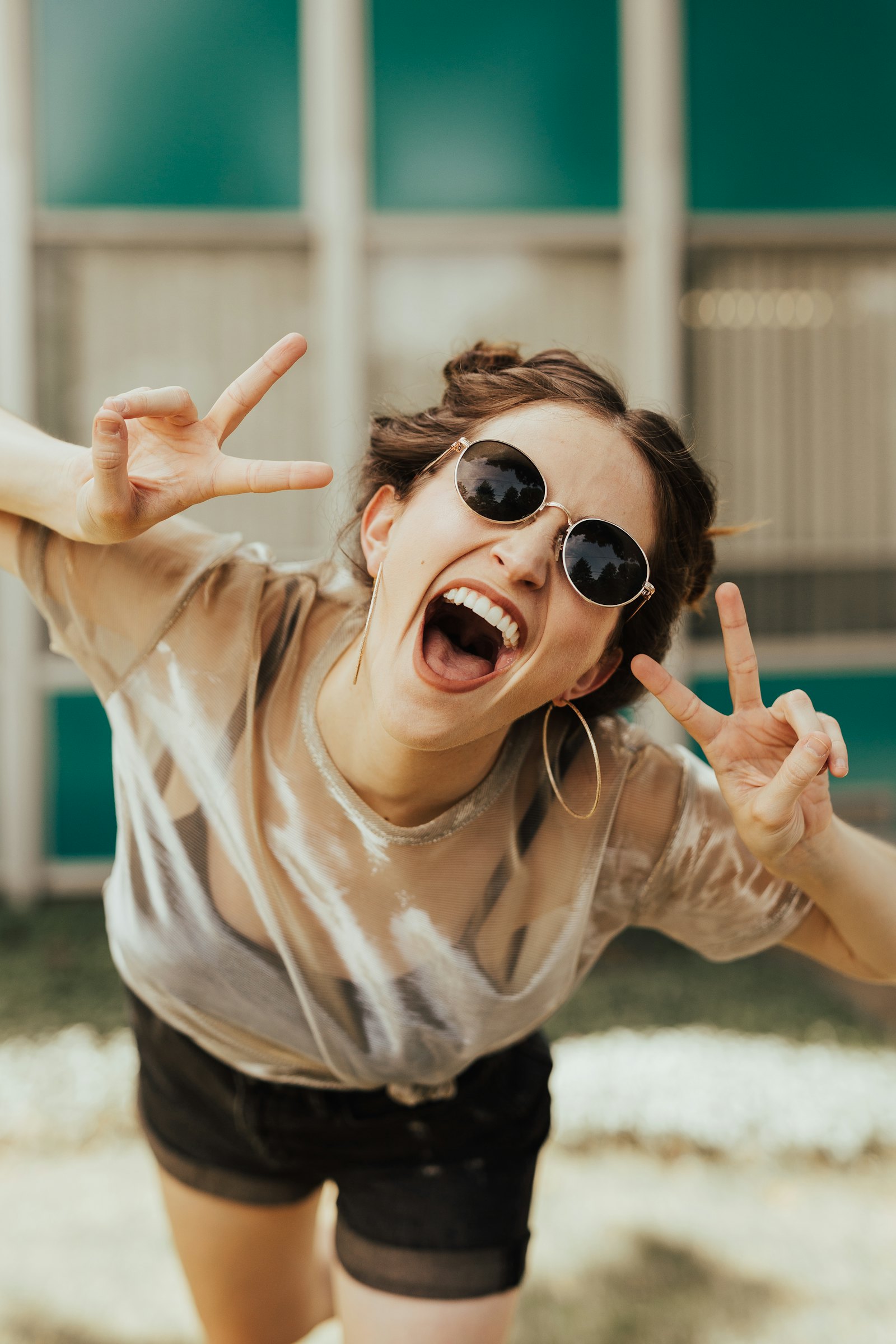A person laughing joyfully in round sunglasses, throwing double peace signs
